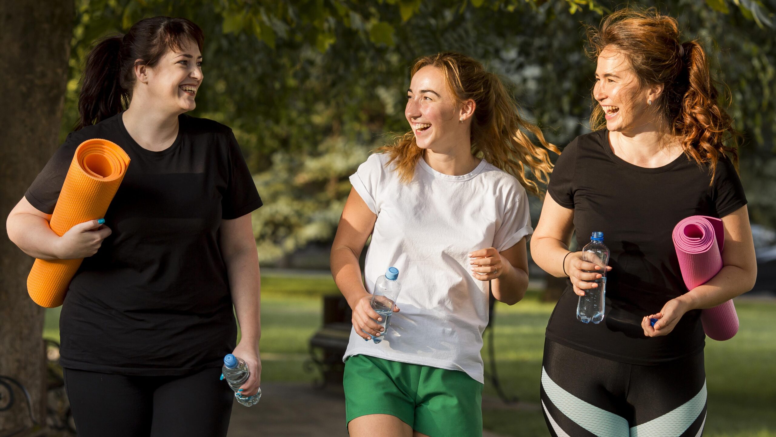 Three women jogging together in a park, smiling and holding water bottles, dressed in athletic wear. The scene captures a lively and healthy outdoor activity, emphasizing fitness and friendship.